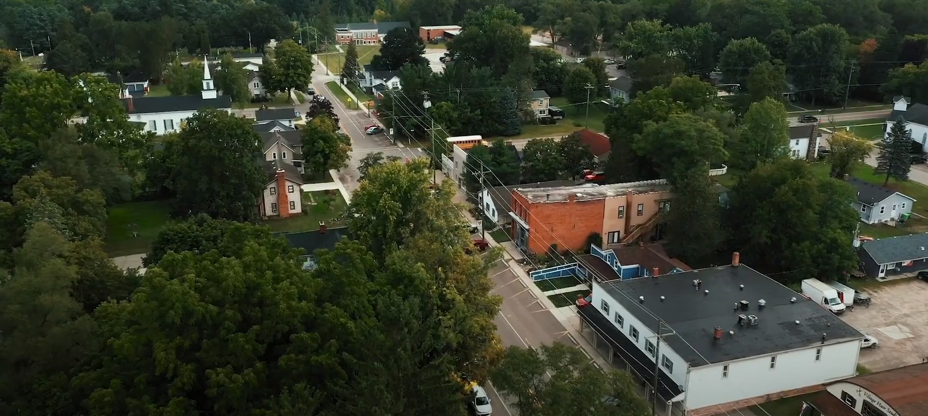 Aerial view of a small town with residential homes, leafy trees, and several roads intersecting, under overcast skies.