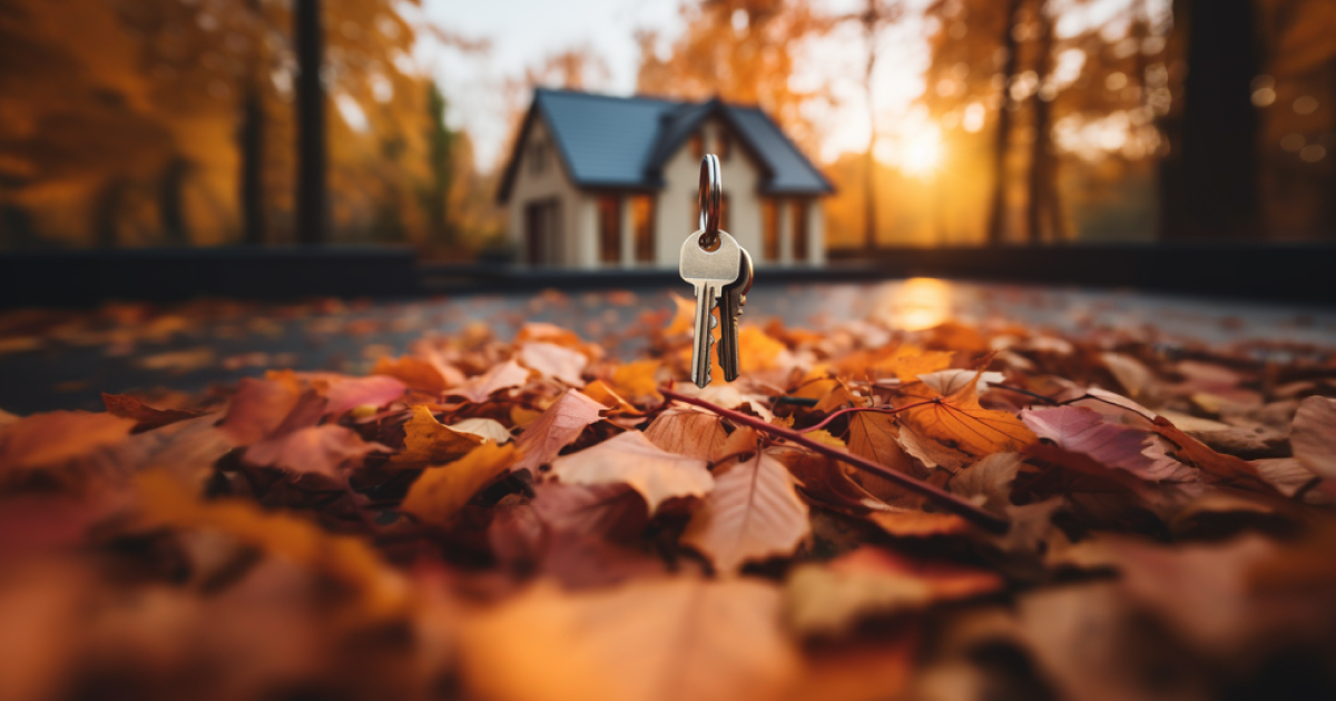 A set of keys hanging above a carpet of colorful autumn leaves, with a blurred house in the background.